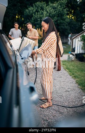 Side view of mature woman charging car with electric plug in back yard Stock Photo