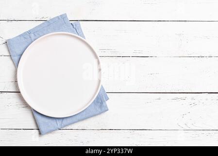 Empty white plate with blue linen napkin on white wooden table, top view, copy space. Food design template background. Stock Photo