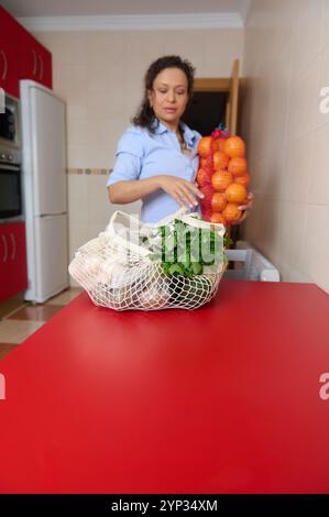 A woman in a kitchen places a bag of oranges on the counter beside a net bag of parsley and white vegetables. The scene conveys a focus on healthy coo Stock Photo