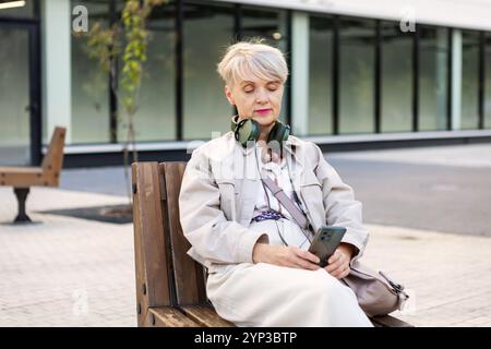 Elegant Mature Woman with Short Hair Wearing Headphones and Using Smartphone While Sitting on Bench Outdoors in City, Active lifestyle for Older adult Stock Photo