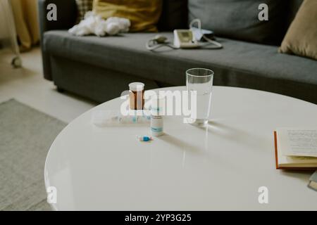 Various medication bottles, glass of water and open book arranged on white table in cozy living room with gray couch in the background Stock Photo