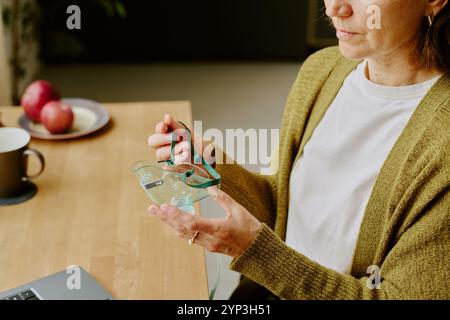 Woman holding her eyeglasses and setting them down on wooden table while sitting, coffee mug and apples arranged near edge of table with laptop in corner Stock Photo