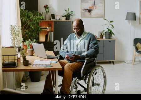 Man in wheelchair using smartphone while working from home office surrounded by books, plants, laptop, and desk setup in a bright, neatly organized room Stock Photo