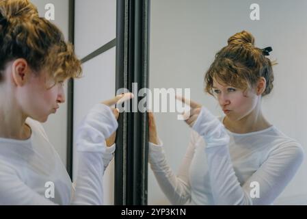 Young woman with curly hair tied in a bun, wearing a white long-sleeved shirt, points at her reflection in a mirror with a displeased expression Stock Photo