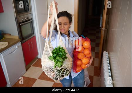 A woman stands in a kitchen, holding a mesh bag with leafy greens and a net bag of oranges. The setting suggests a focus on fresh produce and healthy Stock Photo