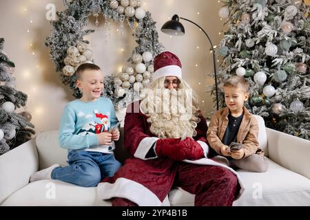 Santa claus sitting on white sofa between two surprised boys and giving them money near decorated christmas tree Stock Photo