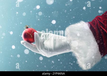 Magical Christmas background with Santa's hand holding a red Christmas decorative ball on a blue background with falling snowflakes Stock Photo
