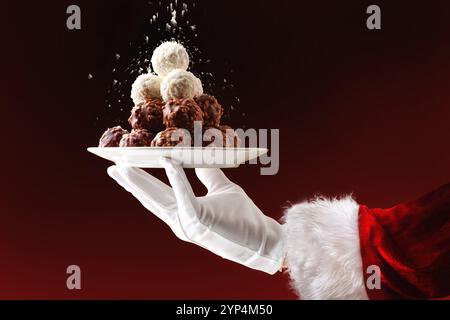 Santa's hand holding plate with pile of coconut and chocolate balls on Christmas with grated coconut shavings falling on top and red and black gradien Stock Photo