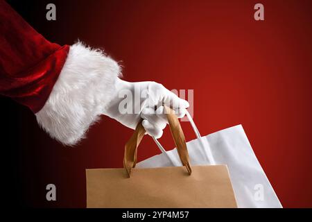 Detail of Santa Claus hand in suit and gloves holding paper shopping bags for shopping on isolated red background. Stock Photo