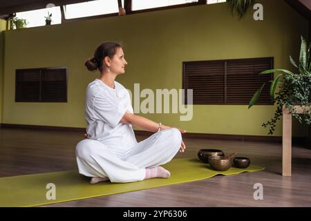 Woman practicing kundalini yoga, doing twisting exercises while sitting in lotus position, practicing in room in white clothes on mat Stock Photo