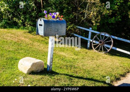 A mailbox with a wheel on the side. The wheel is old and rusty. The mailbox is surrounded by grass and flowers Stock Photo