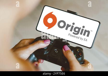 In this photo illustration, the Origin logo is displayed on a smartphone screen as a person plays on a gaming gamepad Stock Photo