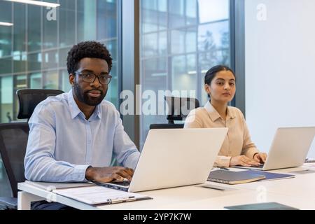 Two professionals engaged in work at laptops in modern office setting. Large windows reflect cityscape, suggesting urban environment. Scene embodies focus, productivity, teamwork, advancement. Stock Photo
