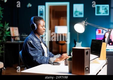 African American woman working on laptop, programming code while listening to music on wireless headset. Female freelancer with headphones using personal computer for monitoring system software. Stock Photo