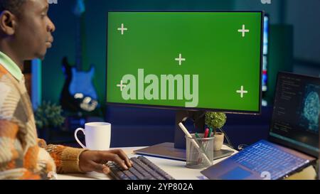 Black person programming neural network code with a green screen on computer to build databases. System engineer working from home while looking at data center security servers. Camera A. Stock Photo