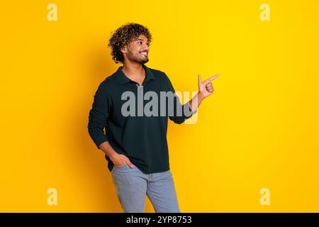 Young man with curly hair pointing to side against vibrant yellow background wearing casual black shirt and jeans for advertising promotions Stock Photo