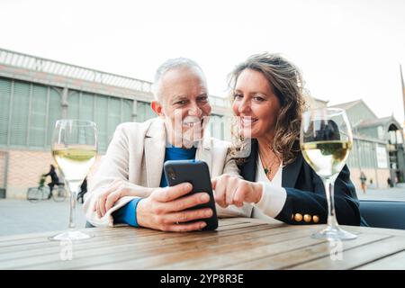 Smiling mature couple enjoying wine together on a cozy outdoor bar terrace, using a smartphone, sharing happy moments, bonding, and relaxing with glasses of white wine on the table. Mid adult people. High quality photo Stock Photo