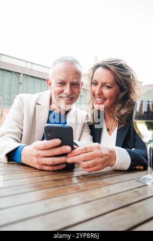 Vertical. Smiling mature couple enjoying wine together on a cozy bar terrace, using a smartphone, sharing happy moments, bonding, and relaxing with glasses of white wine on the table. Mid adult people. High quality photo Stock Photo