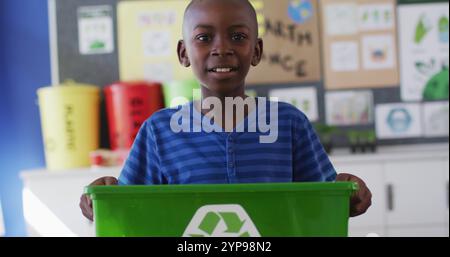 African american schoolboy smiling, holding recycling bin, standing in classroom Stock Photo
