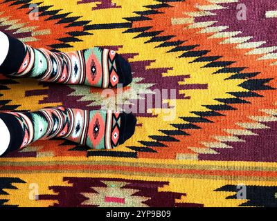 Socks in a geometric pattern. The feet stand on a carpet that has a similar geometric ornament. Different colors. Top view of the feet. Traditional Uk Stock Photo