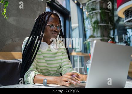 Young black female freelancer with headphones around neck and glasses concentrating on her laptop while working remotely in a trendy cafe, enjoying a refreshing drink during daytime Stock Photo