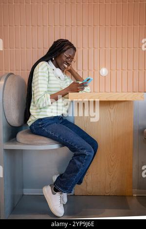 Happy young woman with headphones around her neck using smartphone while sitting at table in modern cafe enjoying break from studying Stock Photo