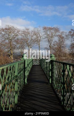 Pretty old footbridge in Kendal, Cumbria with green painted metal lattice work railing with trees in the background under a blue sky with few clouds Stock Photo