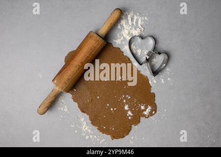 A wooden rolling pin rests on rolled gingerbread dough, alongside a heart-shaped cookie cutter and scattered flour. Stock Photo