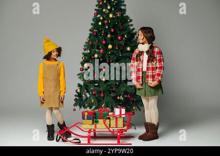 Two cheerful girls share laughter beside a festive Christmas tree with gifts nearby. Stock Photo