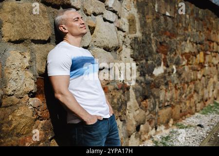 A man in a white shirt and blue jeans is leaning against a stone wall Stock Photo
