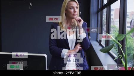 Image of icons and numbers over caucasian businesswoman in office Stock Photo