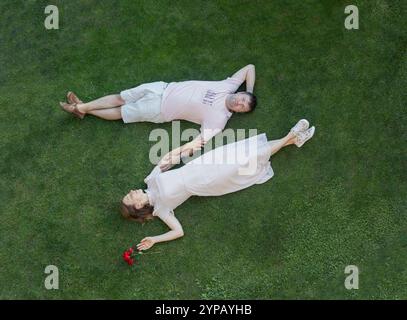 Romantic couple of people over 30 lying on the grass in the park, holding hands. They look happy. Top view. Concept of love, happy relationship. Relax Stock Photo