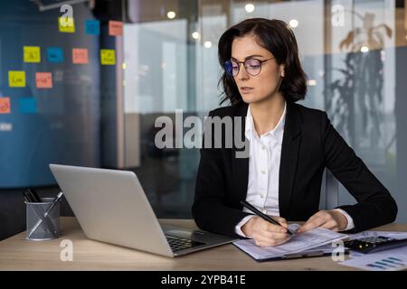 Confident businesswoman in office setting reviewing documents at desk with laptop. Wearing glasses and dark suit, she embodies professional focus, dedication, and modern work environment. Stock Photo