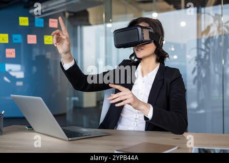 Businesswoman wearing virtual reality headset engages with interactive digital presentation. Utilizing VR technology, she explores immersive workplace solutions at an office desk. Stock Photo