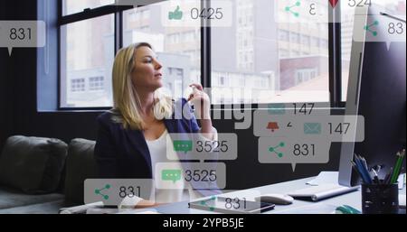 Image of icons and numbers over caucasian businesswoman in office Stock Photo