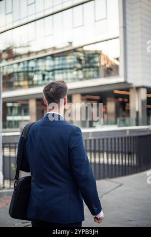 Confident businessman in stylish suit walking near office building in city, carrying laptop bag, looking at modern glass skyscrapers, exuding determin Stock Photo
