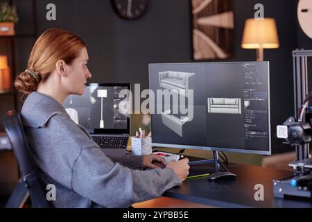 Woman sitting at desk, focusing on 3D modeling software displayed on her computer screen. Office environment includes various technological gadgets and modern decor elements Stock Photo