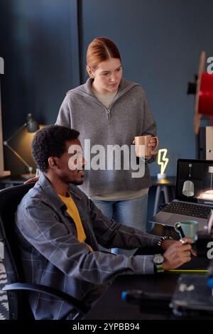 Multiracial colleagues collaborating on project in cozy office space with modern equipment, engaging in focused discussion while reviewing digital content on laptop screen Stock Photo