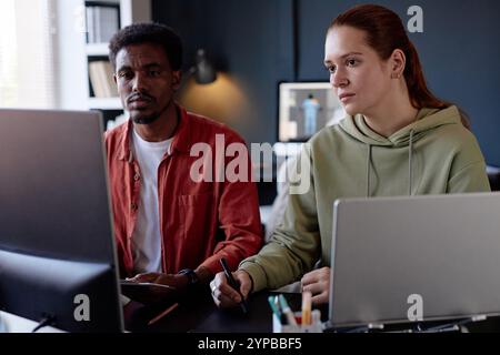 Colleagues focusing on a project using laptops in a modern office with design elements and equipment visible in background, creating a professional and collaborative environment Stock Photo