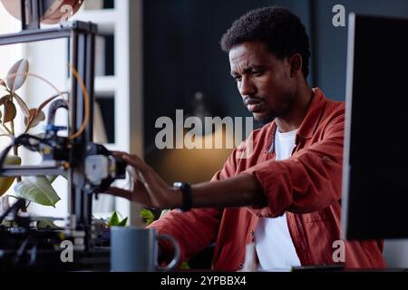Man performing intricate work with a 3D printer, highlighting concentration and precision. The workspace includes plants and various tools emphasizing a creative environment Stock Photo