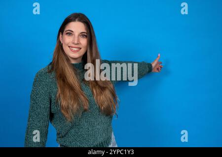 Smiling young woman pointing her finger to the side while standing against a blue background Stock Photo