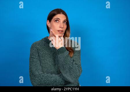 Young woman contemplates with her hand on chin, looking thoughtfully upwards on a vibrant blue backdrop Stock Photo