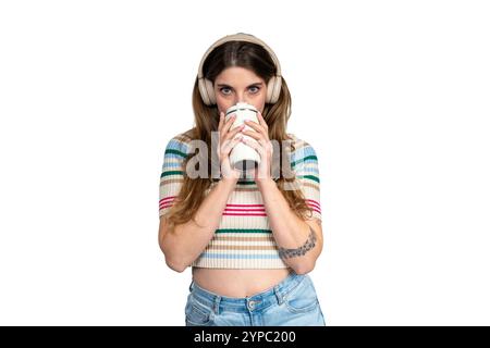 Young woman enjoying a hot beverage in a reusable cup while wearing headphones and listening to music on a white background Stock Photo