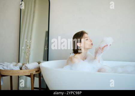 Caucasian brunette woman blowing foam in air while taking relaxing hot bathtub at home Stock Photo