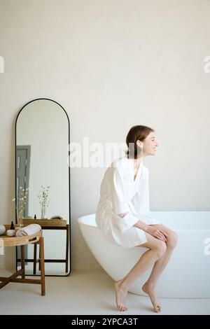 Vertical shot of young adult woman in white bathrobe sitting on bath and resting after work week Stock Photo