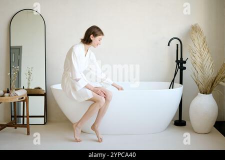Young smiling lady dressed in silk bathrobe sitting on bathtub and touching water to check its temperature Stock Photo