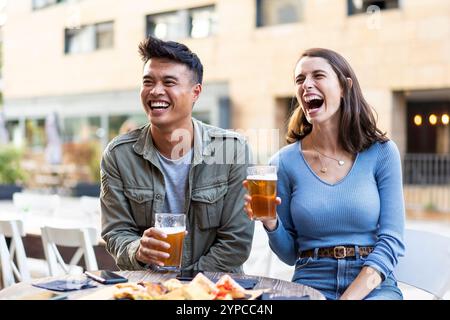 Happy friends laughing and enjoying beers at an outdoor cafe. Young adults socializing and having fun over drinks Stock Photo