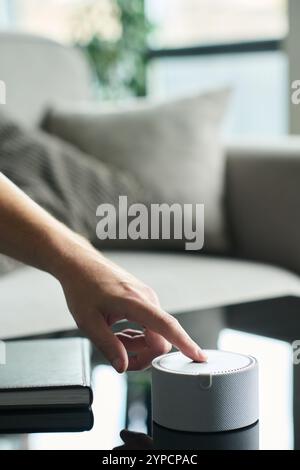 Hand reaching to turn on smart speaker on glass coffee table in bright, modern living room featuring cozy furnishing and natural light streaming through windows Stock Photo