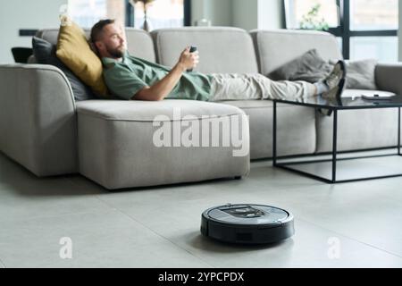 Man lying on a couch using a smartphone while a robot vacuum is cleaning the floor nearby. Modern living room setup with large windows and minimalist furniture Stock Photo
