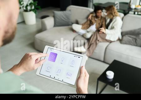 Individual using tablet for controlling smart home features while couple sits on sofa in background. Tablet displays various application icons for home automation Stock Photo